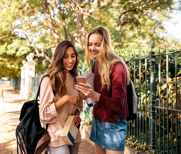 a student shows her phone to one of her friends while on a wooded sidewalk